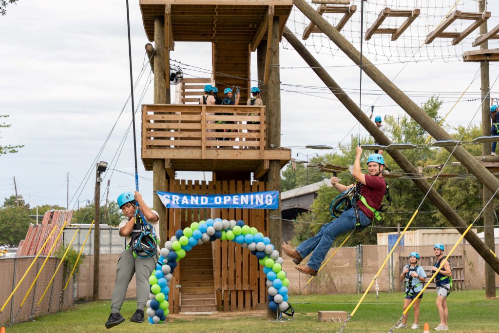 Two male college students on a zip line with a challenge course in the background.