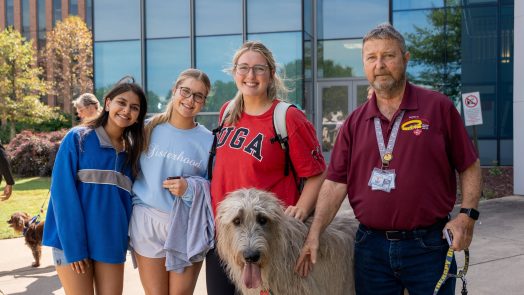 Three college-aged female students pose for a photo with a man and a therapy dog.