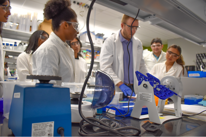 A group of five college students gather around a male professor in a scientific lab as he explains how an experiment should go.