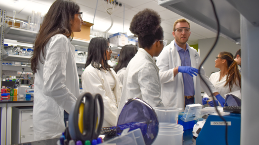 A male college professor speaks with a group of college students in a scientific lab.