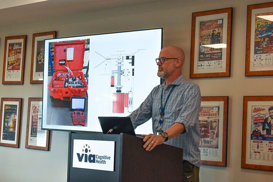 A man stands at a podium at the front of a classroom and delivers a presentation to a group of people.