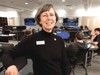 A female computer sciences professor sits in a chair in a classroom and smiles at the camera.