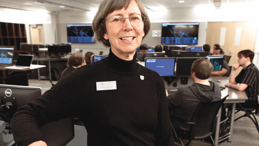 A female computer sciences professor sits in a chair in a classroom and smiles at the camera.