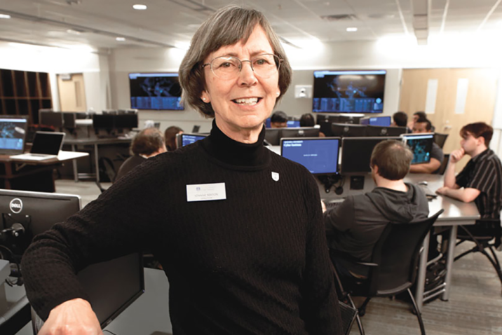 A female computer sciences professor sits in a chair in a classroom and smiles at the camera.