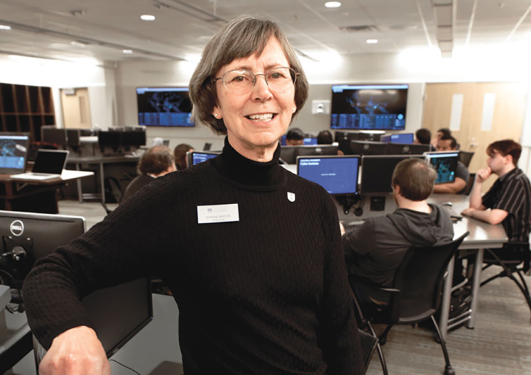 A female computer sciences professor sits in a chair in a classroom and smiles at the camera.