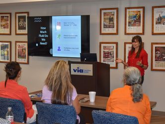 A woman stands at the front of a room in front of a group of people while delivering a presentation.