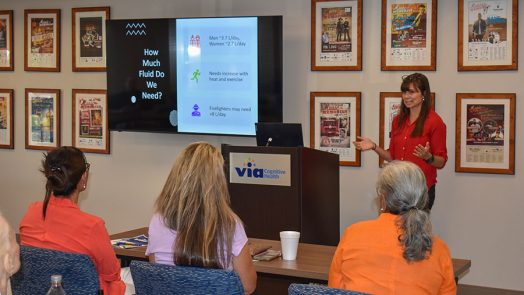 A woman stands at the front of a room in front of a group of people while delivering a presentation.
