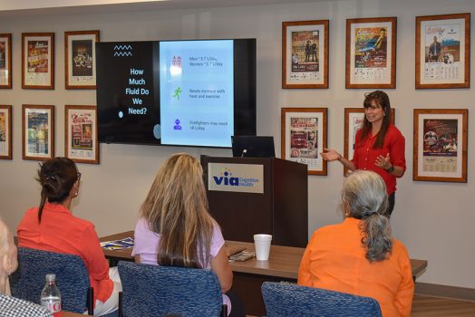 A woman stands at the front of a room in front of a group of people while delivering a presentation.