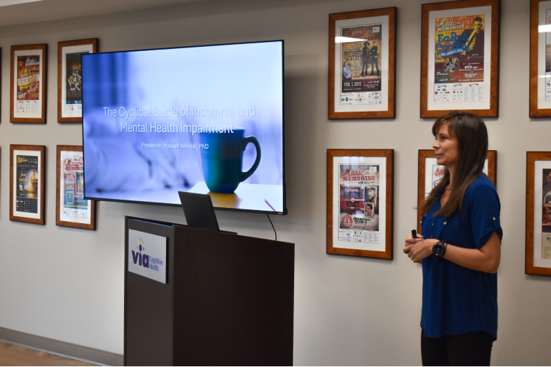 A woman stands at the front of a room in front of a group of people while delivering a presentation.