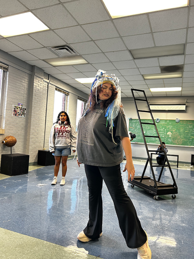 Woman in headdress poses with closed eyes and a ladder and another woman in background.