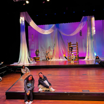 Two women sit on a stage with a spread out group of people behind them, all in front of a large backdrop.