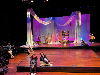 Two women sit on a stage with a spread out group of people behind them, all in front of a large backdrop.