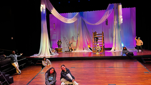 Two women sit on a stage with a spread out group of people behind them, all in front of a large backdrop.