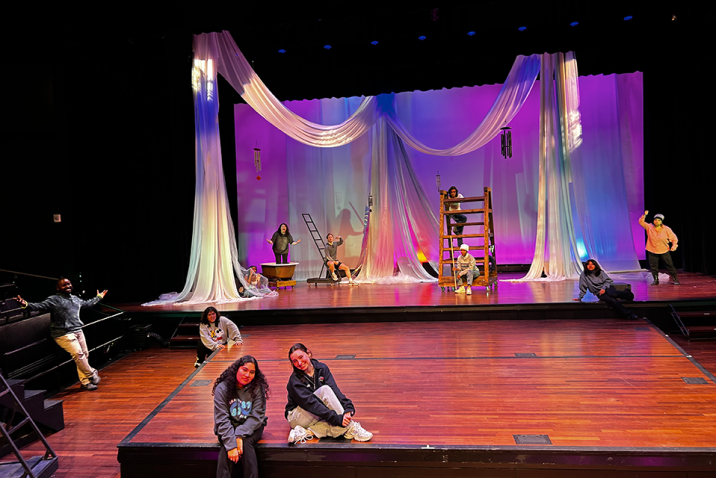 Two women sit on a stage with a spread out group of people behind them, all in front of a large backdrop.