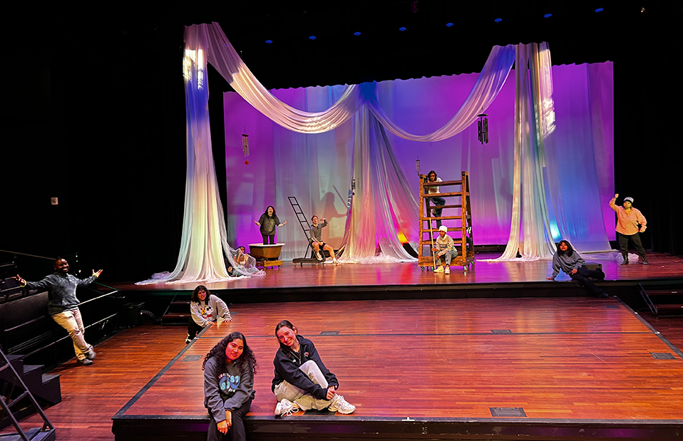 Two women sit on a stage with a spread out group of people behind them, all in front of a large backdrop.