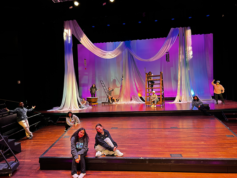 Two women sit on a stage with a spread out group of people behind them, all in front of a large backdrop.