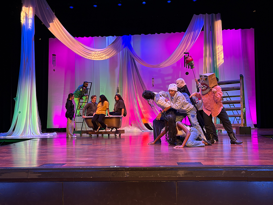 Ten people on a stage in various acting poses with large props behind them.
