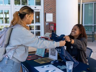 A woman hands another woman a t-shirt at a table outside during a special event.