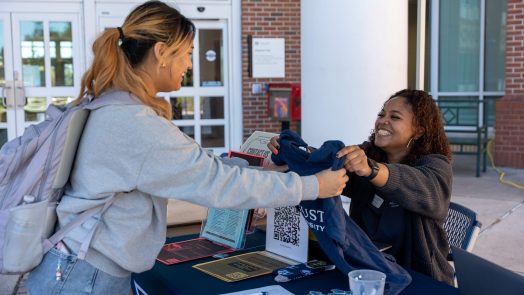 A woman hands another woman a t-shirt at a table outside during a special event.