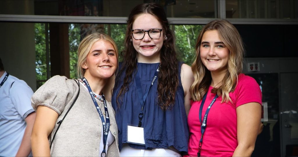 Three female high school students smile while waiting for an event on a college campus to begin.