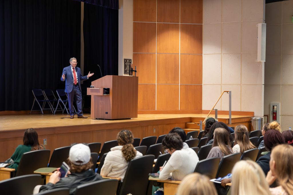 A man stands on a stage near a podium and delivers a lecture to a group of college students in a large auditorium.