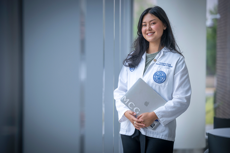 A female medical student wearing a lab coat and holding a laptop smiles at the camera.
