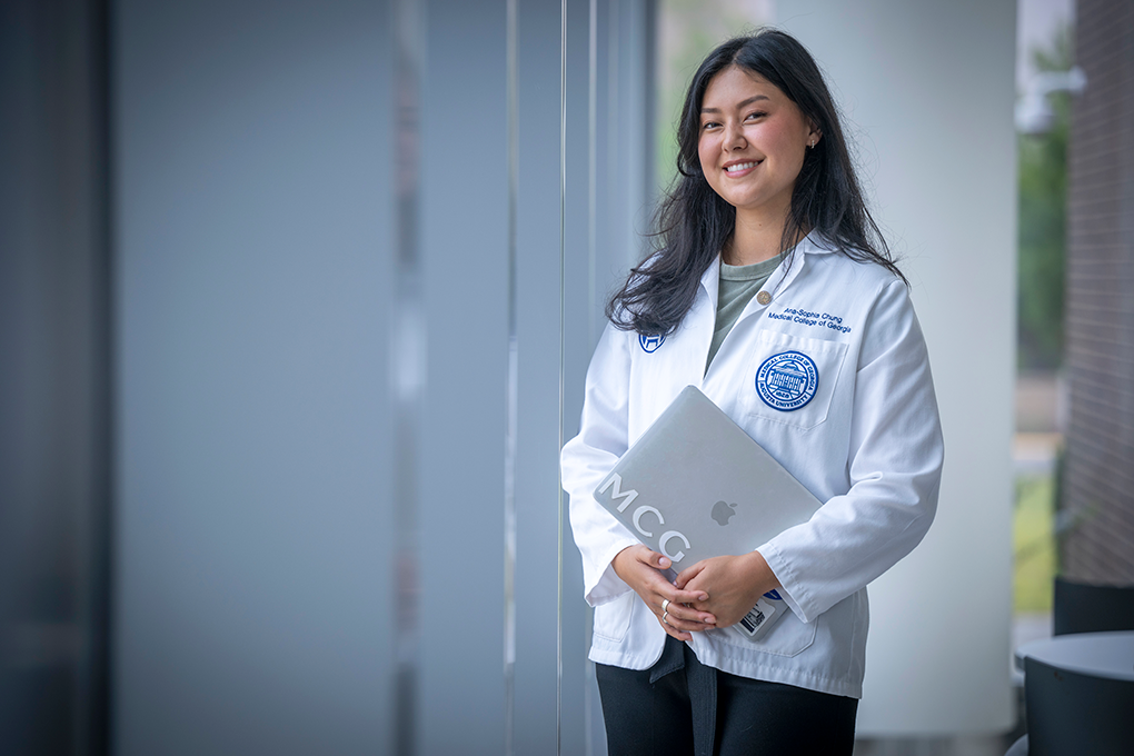 A female medical student wearing a lab coat and holding a laptop smiles at the camera.