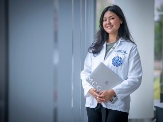 A female medical student wearing a lab coat and holding a laptop smiles at the camera.