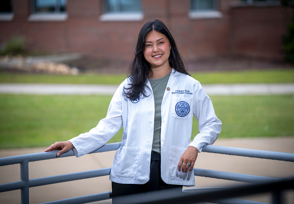 A female medical student wearing her lab coat leans against an outdoor railing.
