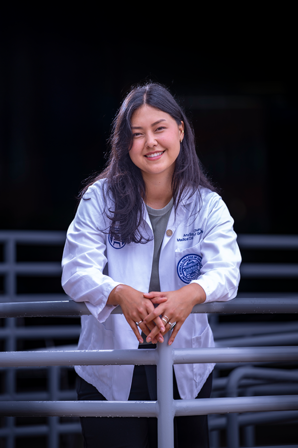 A female medical student wearing her lab coat leans against an outdoor railing.