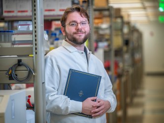 A male college student wearing a scientific lab coat and holding a folder stands in a large science lab.