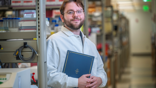 A male college student wearing a scientific lab coat and holding a folder stands in a large science lab.