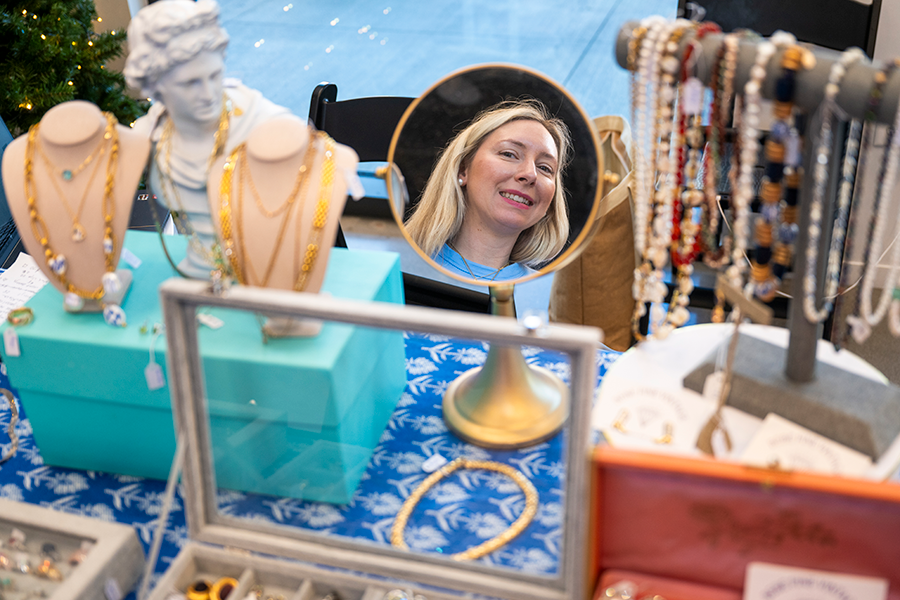 A woman looks into a mirror and smiles. There is jewelry spread out on a table around the mirror.