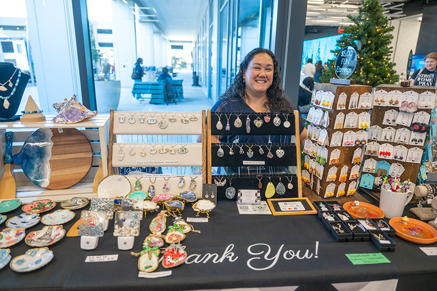 A woman stands behind a table that has lots of handmade jewelry on stands. The woman is taking part in a small business market.