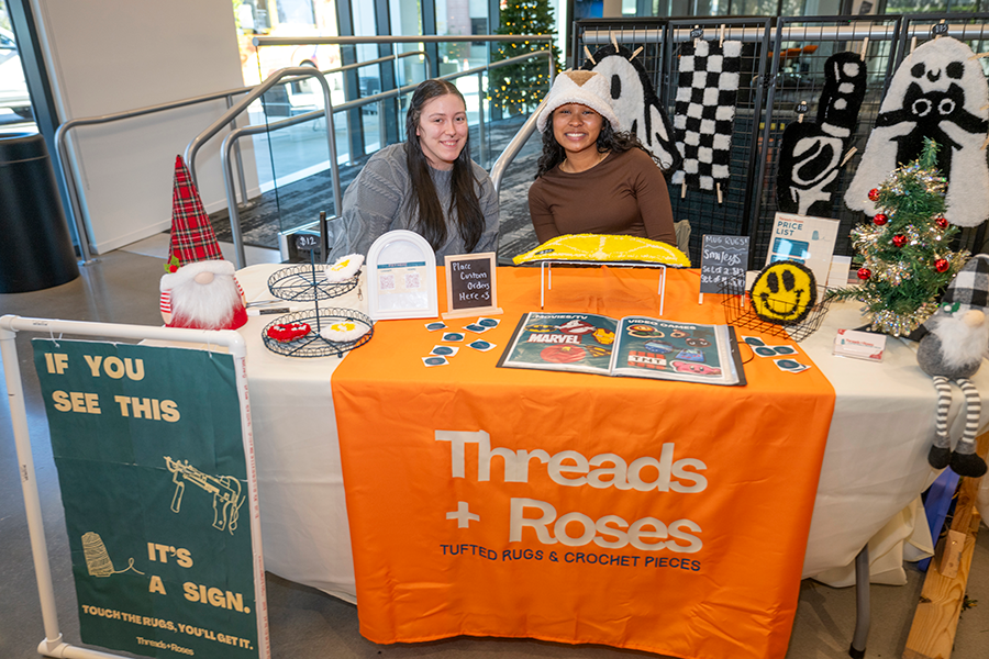 Two college-aged women sit at a booth during a small business market. They are selling hand-made rugs.