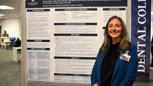 Woman in a blazer smiles in front of a poster project, she is in a library