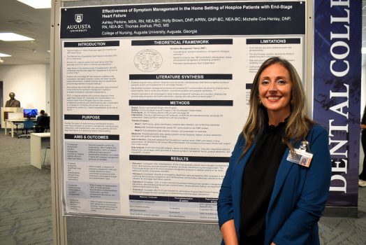 Woman in a blazer smiles in front of a poster project, she is in a library