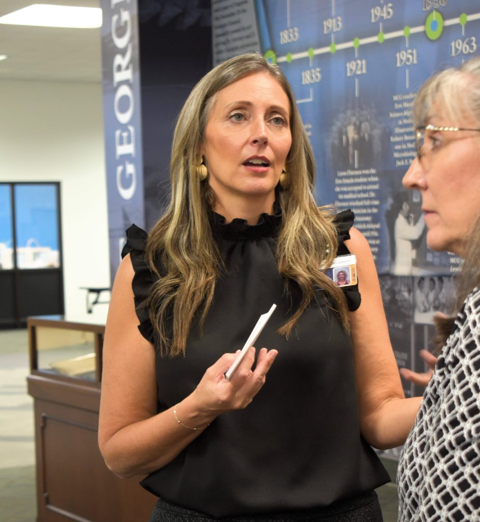 A woman, with notecards in her hand, stands in a library and speaks to another woman