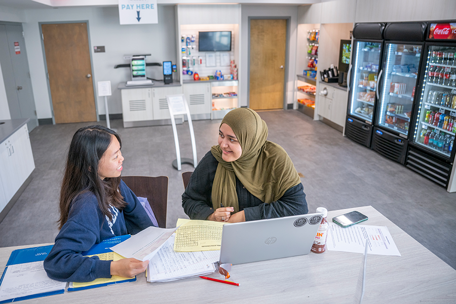 Two college-aged women sit at a table inside a small cafeteria inside a college library.