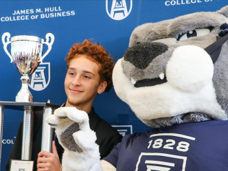 A male high school student holds a large trophy and stands next to a college mascot depicting a large cat.