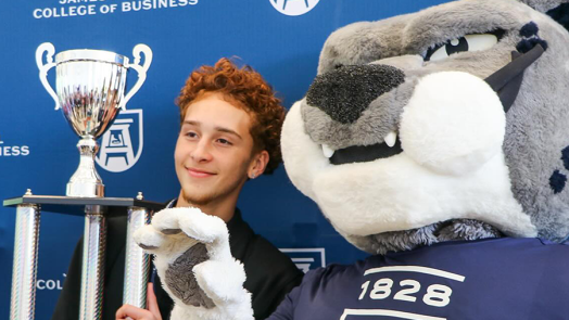 A male high school student holds a large trophy and stands next to a college mascot depicting a large cat.