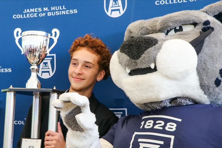 A male high school student holds a large trophy and stands next to a college mascot depicting a large cat.