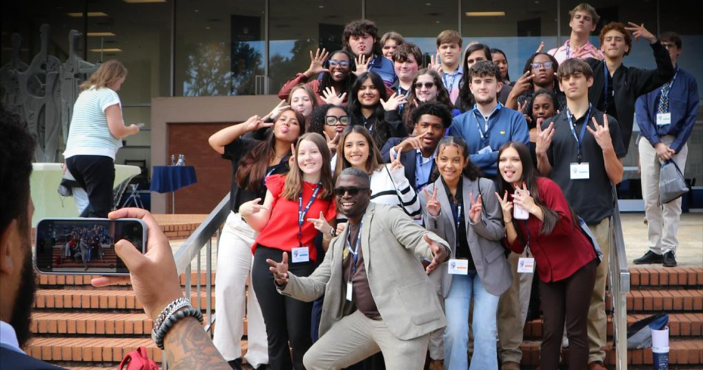 A group of high school students pose for a picture on the outside steps of a building on a university campus.
