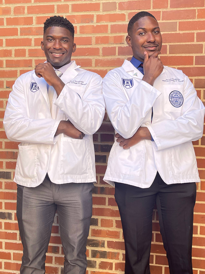 Two male medical students who are brothers wear scientific lab coats that say 'Medical College of Georgia' and pose for a picture.