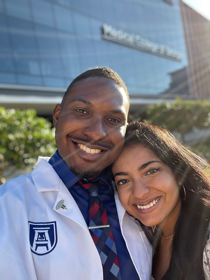 A man wearing a scientific lab coat and his fiancee take a selfie in front of a modern medical college building.