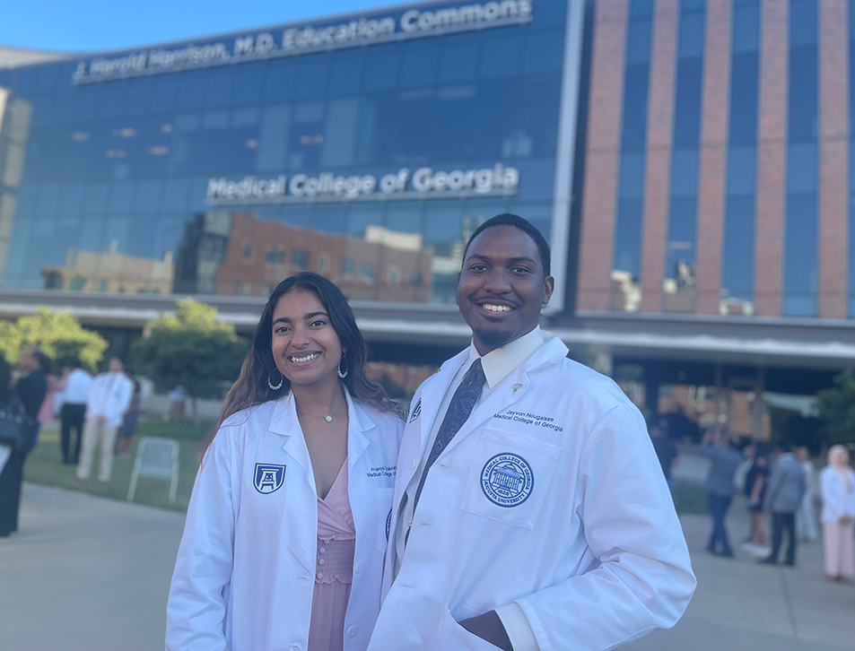 A female medical student and a male medical student wearing lab coats pose for a photo in front of a modern medical school building.