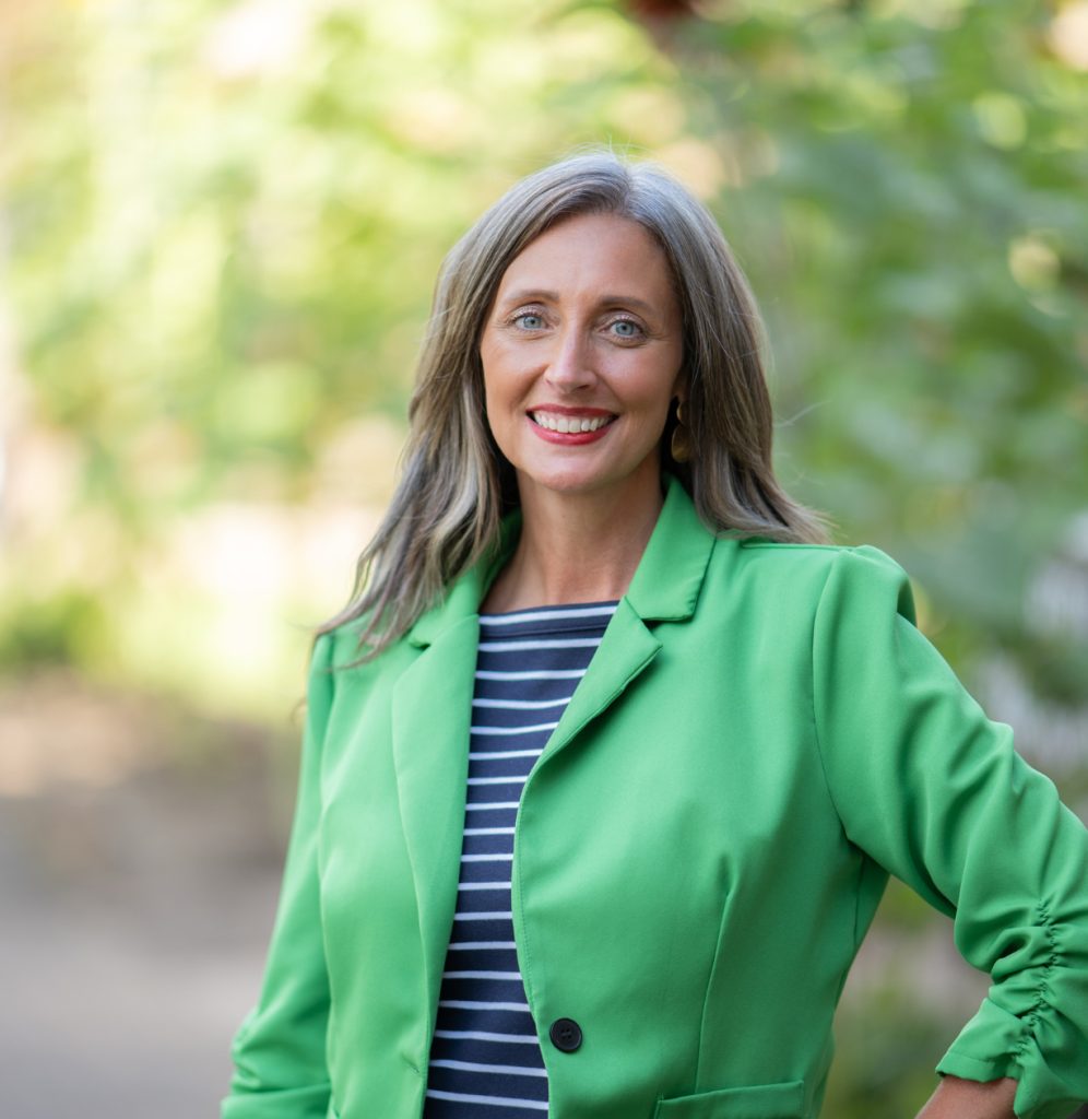Woman in a blazer poses for a professional headshot, a blurred out background of trees and greenery is behind her