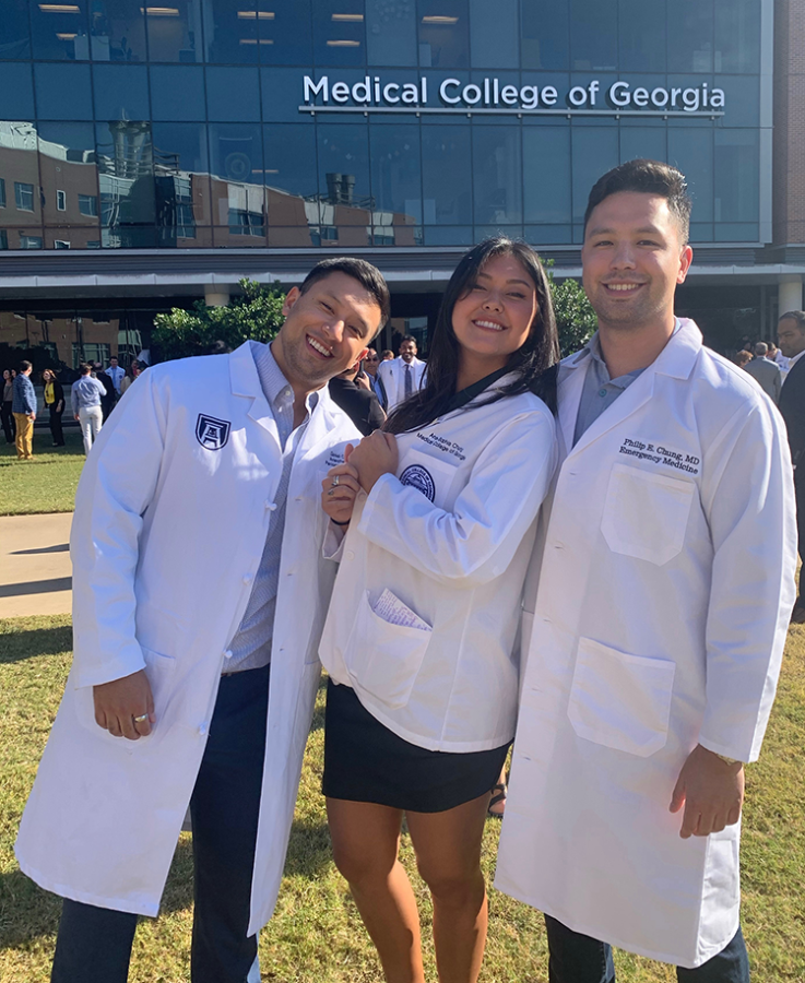 Three siblings, including two men and one woman, pose for a photo outside. All three are wearing scientific lab coats.