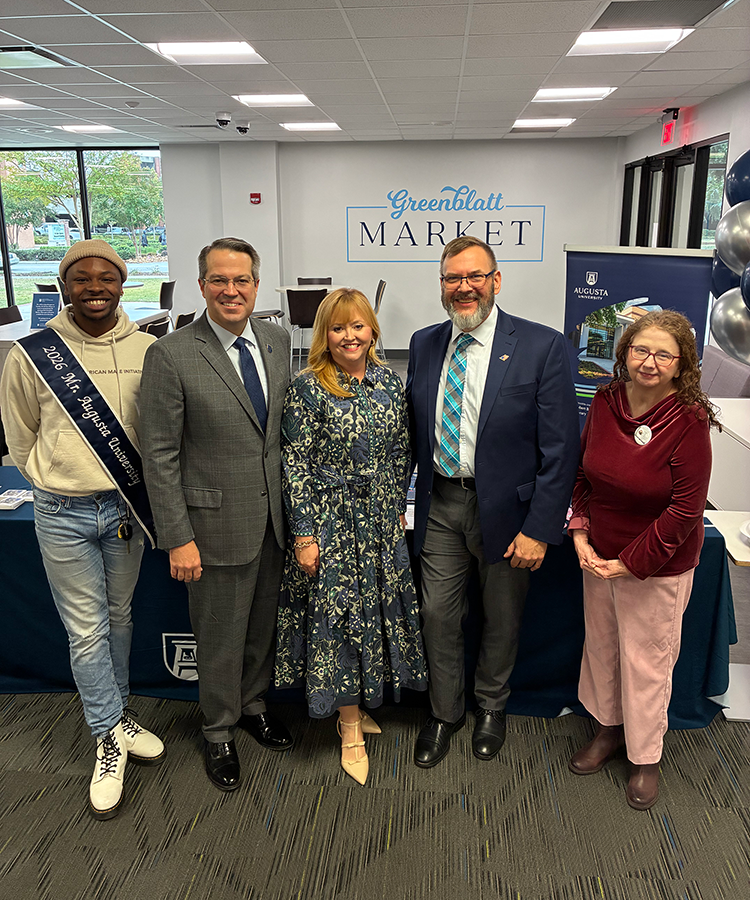 Two men and two women stand with a male college student during a special ribbon-cutting event on a college campus inside a library.
