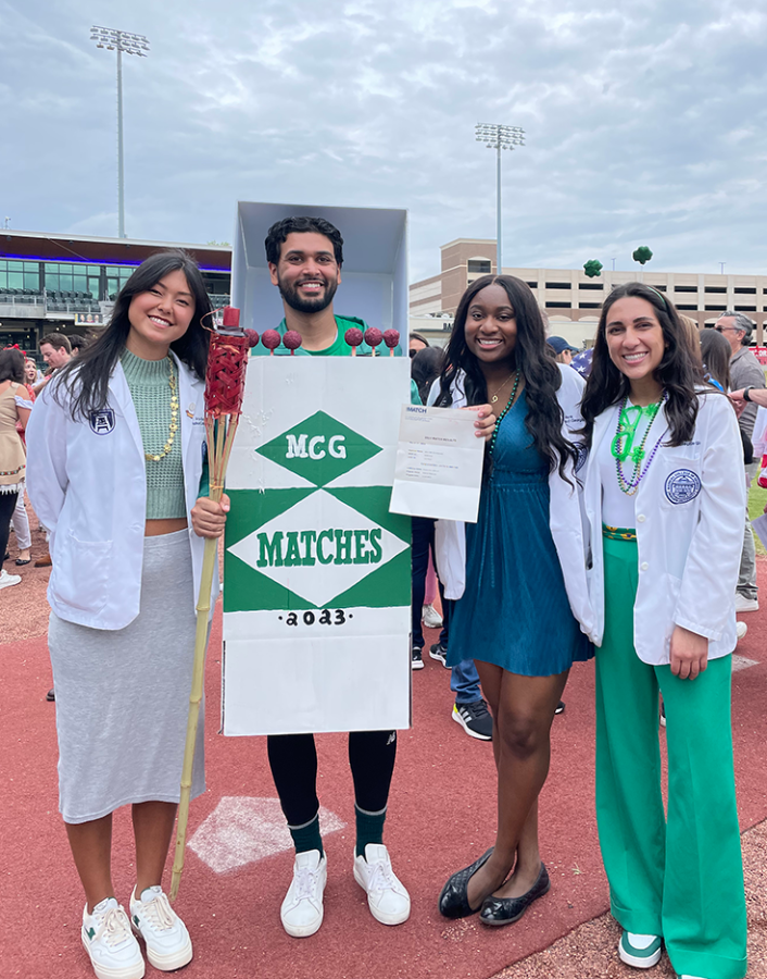 Four medical students stand together during an outdoor ceremony. Three of the students, all women, are wearing their scientific lab coats while the man is dressed up as a box of oversized matches, since the celebration is called Match Day.